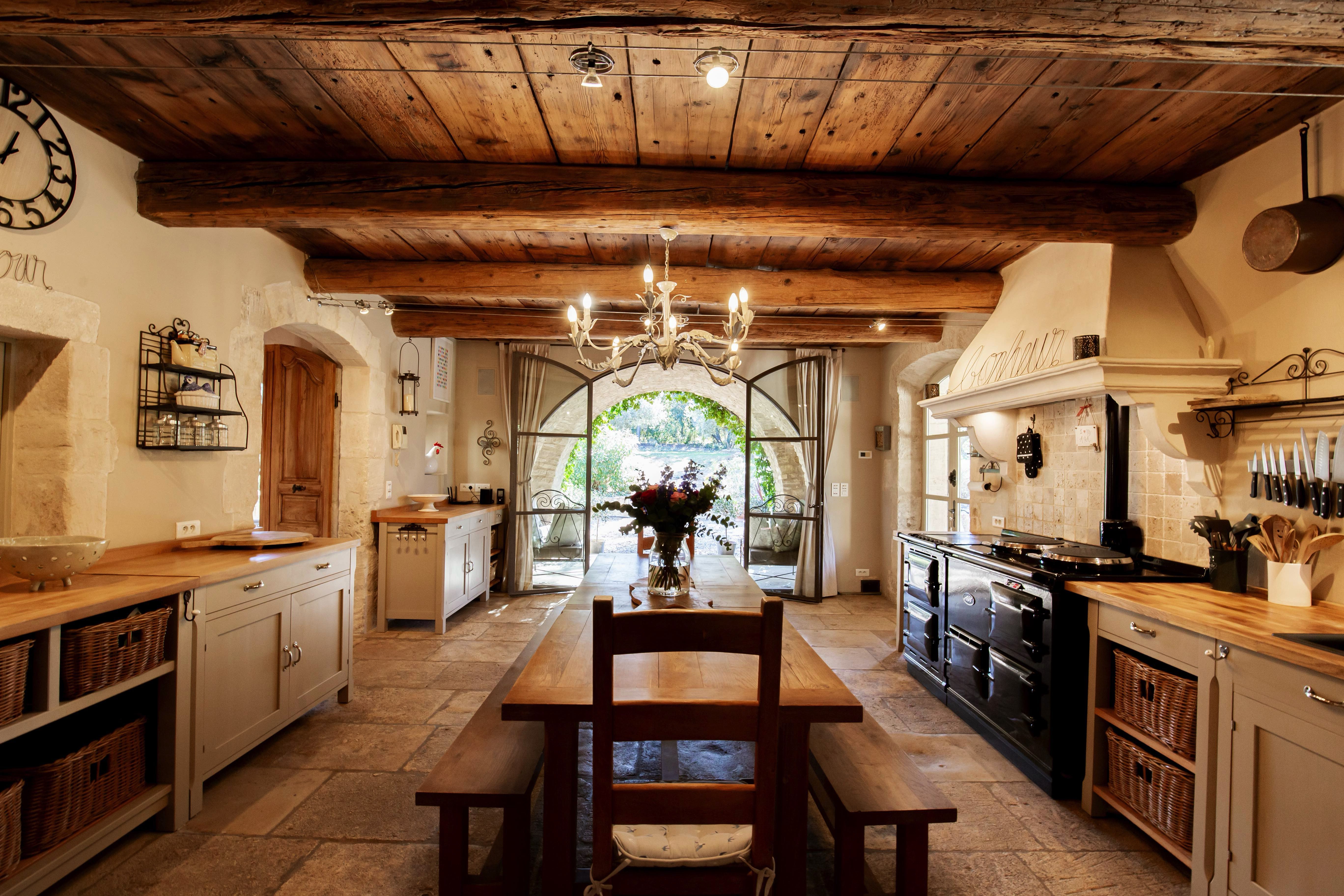 A spacious, rustic kitchen featuring wooden beams, stone walls, a large chandelier, and a central dining table. arched doors open to the outside at the far end.